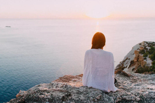 Woman in white cover-up sitting on rocky cliff facing the sea at sunset