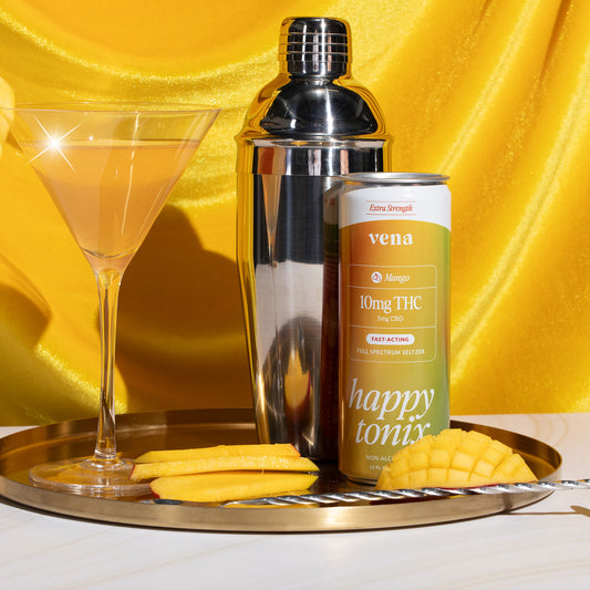 Front view of Happy Tonix Mango 10mg THC seltzer can showing ‘10mg THC’ label with martini glass, cocktail shaker, sliced mango on yellow satin backdrop