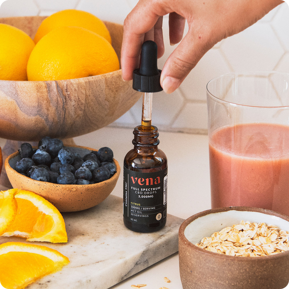 Hand holding dropper above Vena Full Spectrum CBD citrus tincture bottle showcasing amber dropper with oranges, blueberries, glass of juice, and oats