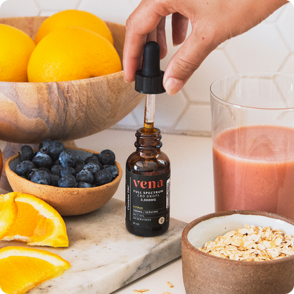 Hand holding dropper above Vena Full Spectrum CBD citrus tincture bottle showcasing amber dropper with oranges, blueberries, glass of juice, and oats