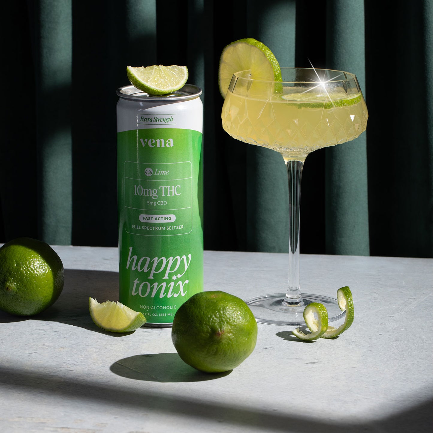 Front view of Happy Tonix Lime 10mg THC seltzer can showing ‘10mg THC’ label beside a cocktail glass and fresh limes on a sunlit tabletop