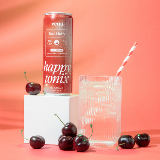 Front view of Vena Happy Tonix Black Cherry seltzer can showing label text next to a ribbed glass with straw and fresh cherries on a pink background