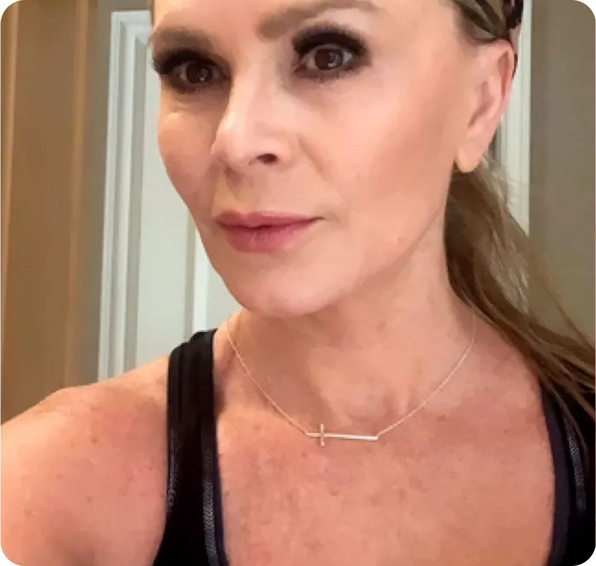 Close-up head-and-shoulders selfie of a woman wearing a dark tank top and a horizontal cross necklace, hair pulled back, neutral makeup.