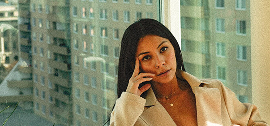 Young woman by a window wearing a beige coat and necklaces, looking at the camera with city buildings outside.