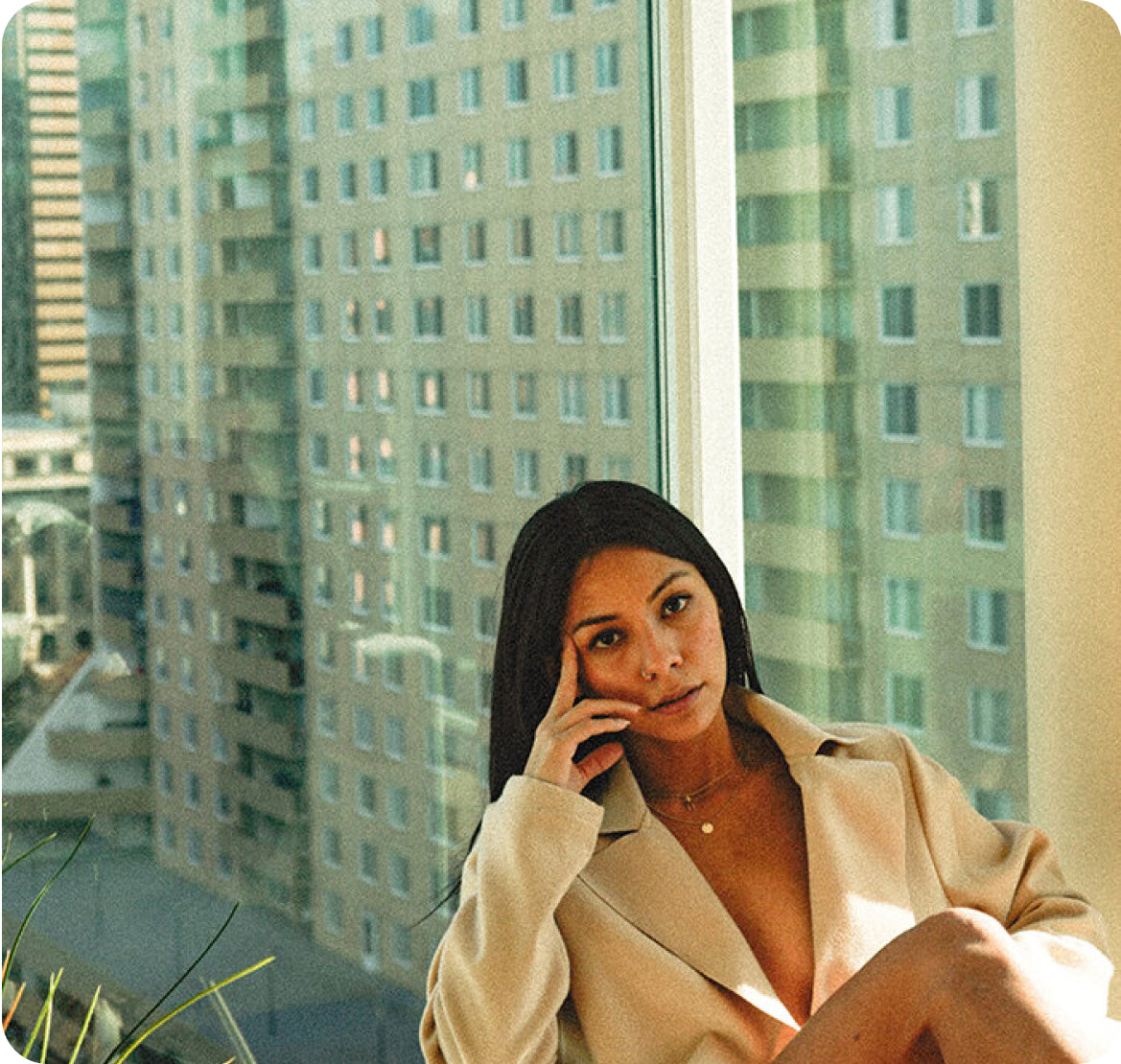 Woman in a beige blazer seated by a high‑rise window with city buildings outside, thoughtful expression