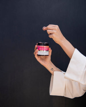 Close-up of No Worries gummy jar held by two hands with a coin about to drop into the open jar, pink label and small heart wrist tattoo against dark background