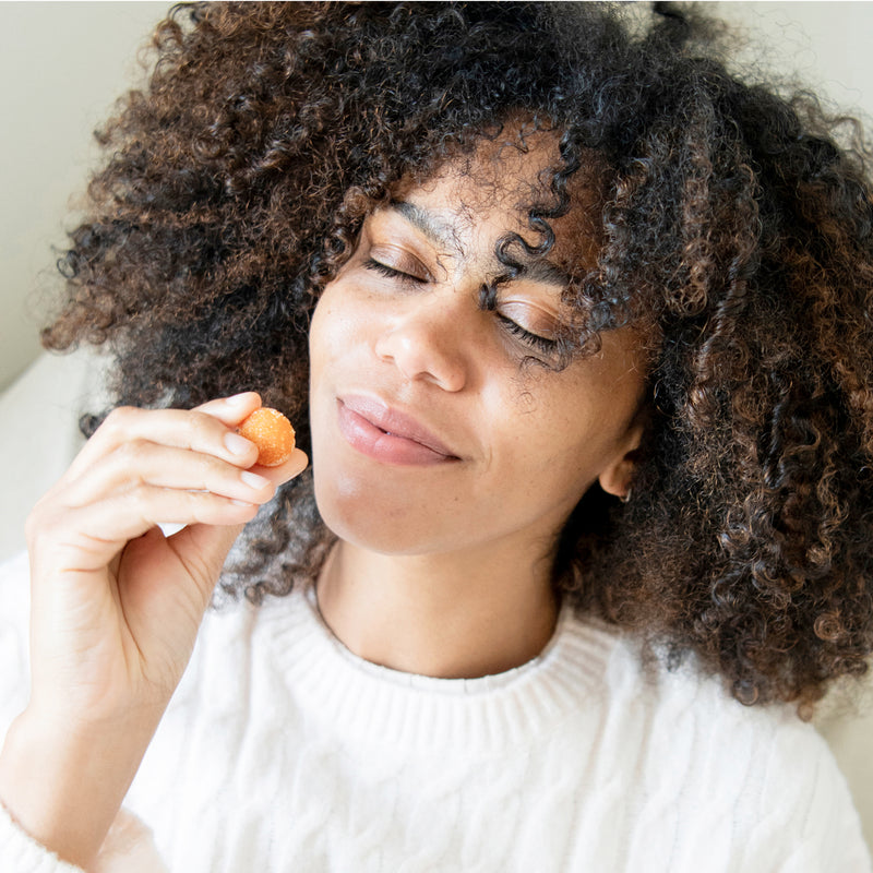 Smiling woman with curly hair holding a small orange gummy near her mouth, eyes closed, savoring the candy.