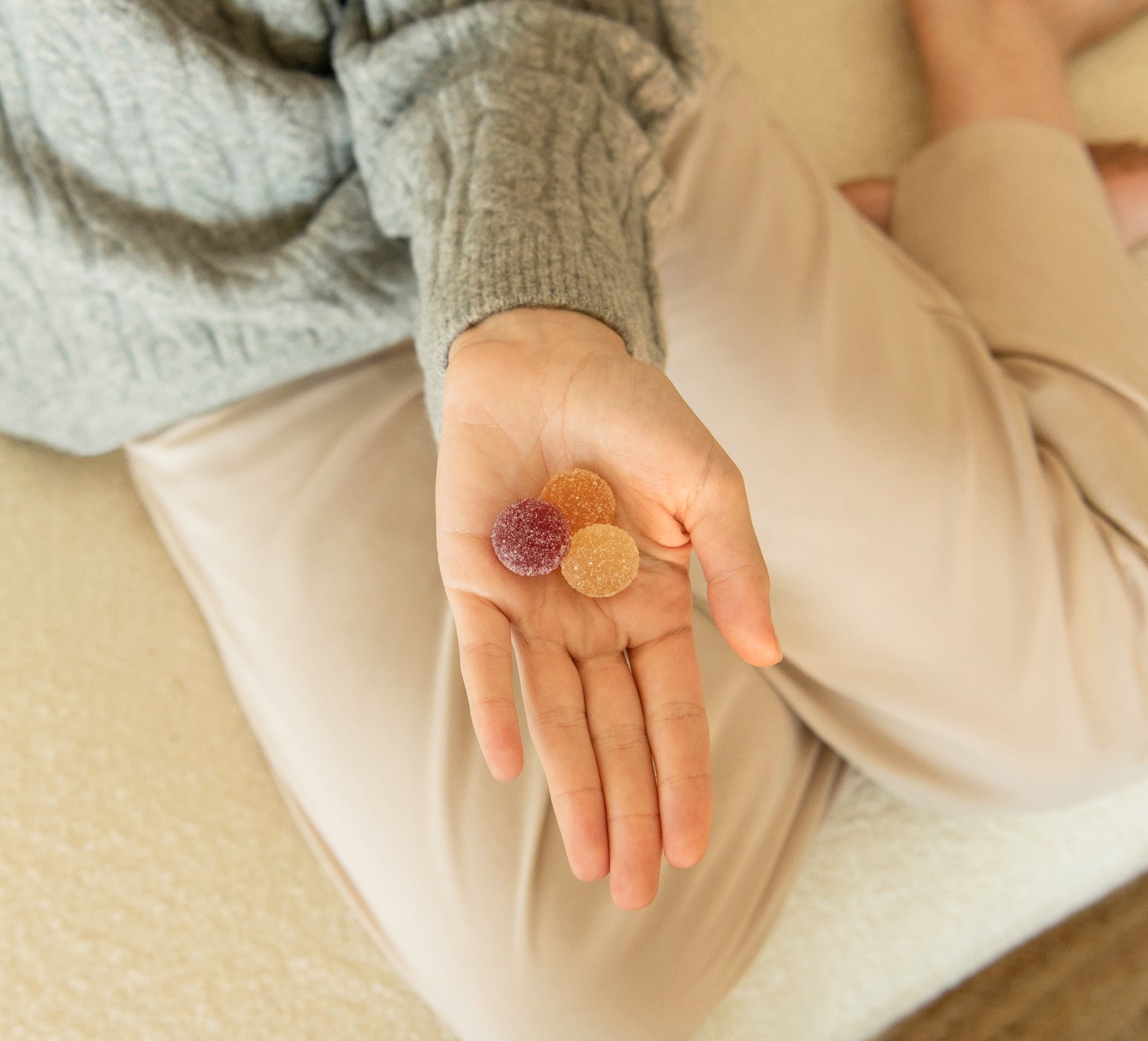 Person's open hand holding three sugar-coated fruit gummies (one purple, two orange) resting on their lap
