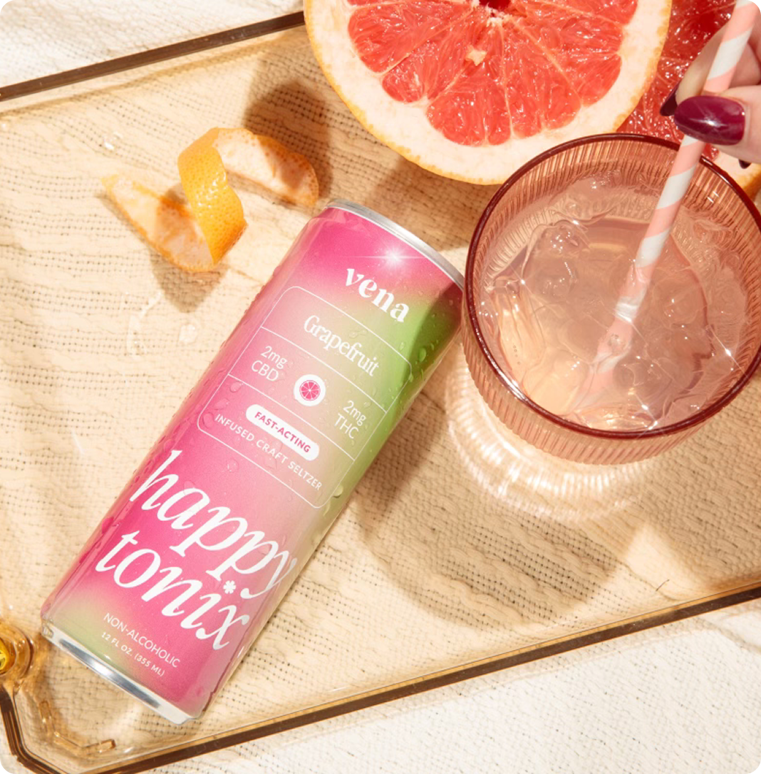 Tilted Happy Tonix Grapefruit seltzer can with pink to green artwork and branding beside grapefruit half and glass on tray