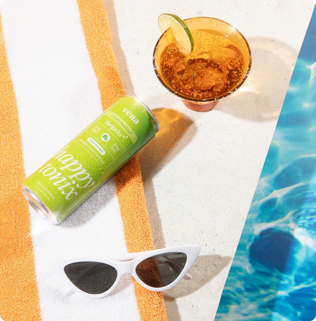 Green can reading 'happy tonix', 'vena' and 'THC' on towel beside cocktail with lime, white sunglasses and pool edge.