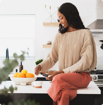 Woman sitting on a kitchen counter holding a CBD tincture bottle and preparing a cup