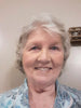Smiling older woman with short white hair wearing a blue patterned blouse against a neutral wall.