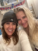 Two smiling women wearing winter hats and sweaters posing inside a store with clothing racks behind them.