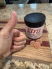 Jar of pink body scrub on a wooden cutting board with a hand giving a thumbs-up beside it.