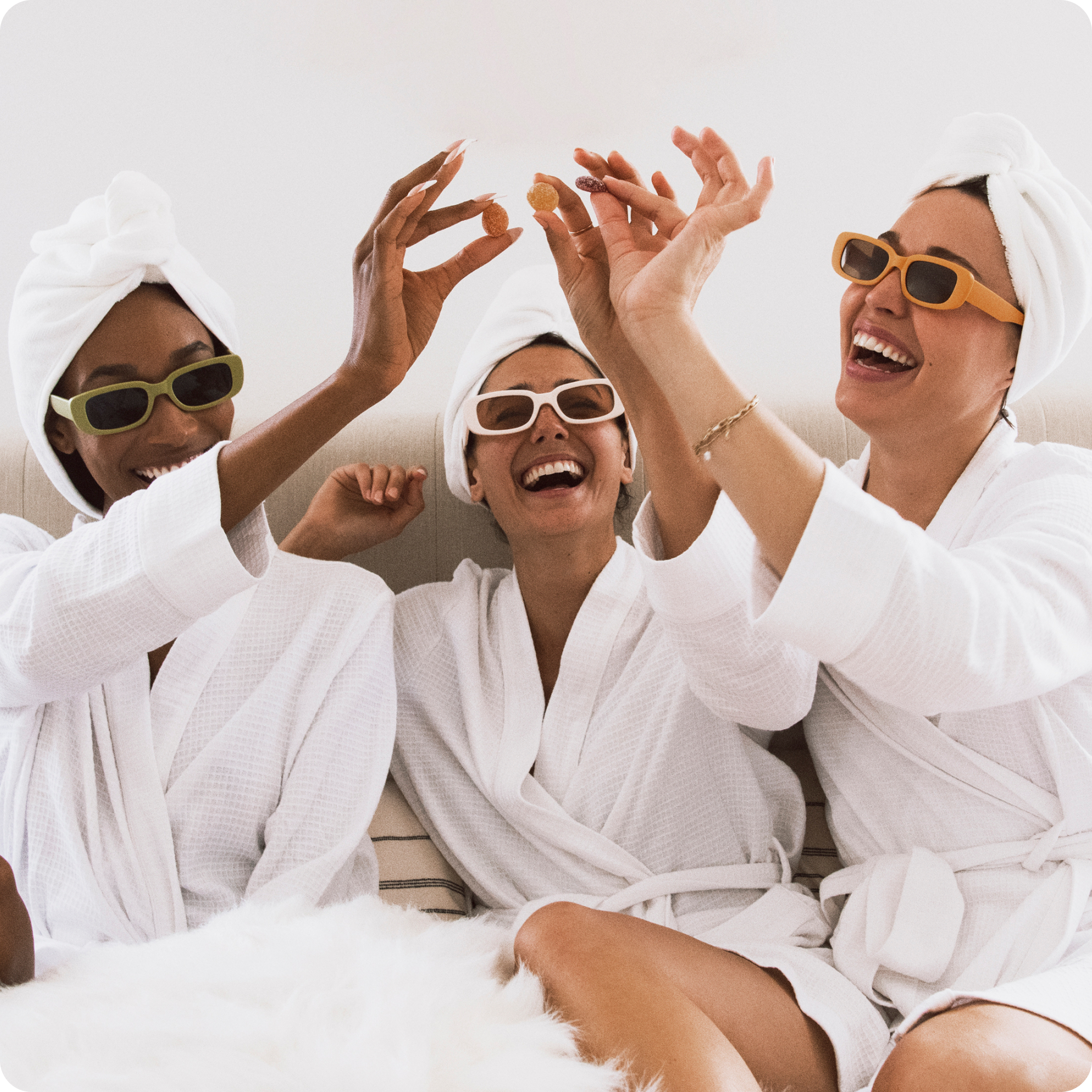 Three friends in white spa robes and towels on their heads, wearing sunglasses and laughing while holding round candies.